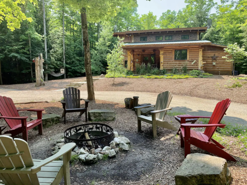 A cozy log cabin surrounded by trees, with a fire pit and colorful chairs arranged in a circle in the foreground.