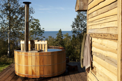 A wooden hot tub on a deck with a scenic view of the ocean and trees under a clear blue sky.