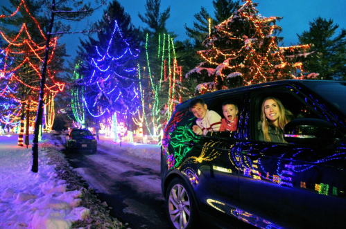 A family enjoys colorful holiday lights from their car, surrounded by snow-covered trees at night.