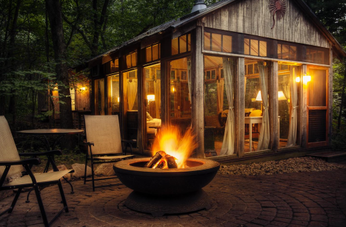 A cozy cabin with large windows, illuminated by warm lights, beside a fire pit surrounded by chairs in a wooded area.