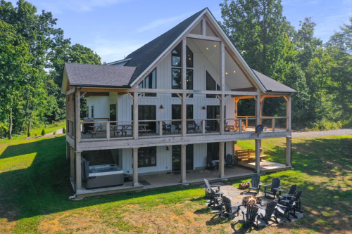 A modern two-story house with a large deck, surrounded by trees and a fire pit area in the foreground.