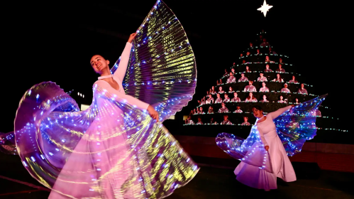 Two dancers in flowing white dresses twirl with illuminated, colorful wings against a backdrop of a starry Christmas tree.
