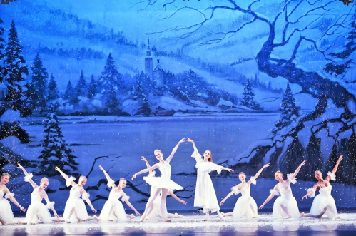 A group of ballet dancers in white costumes perform on stage against a snowy winter backdrop.