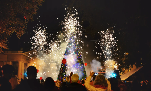 A crowd gathers around a brightly lit Christmas tree, surrounded by fireworks and festive decorations at night.