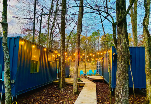 Two blue shipping containers surrounded by trees, connected by a wooden path and adorned with string lights.