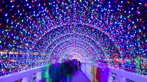 A vibrant tunnel of colorful lights with blurred figures walking through, creating a festive atmosphere.