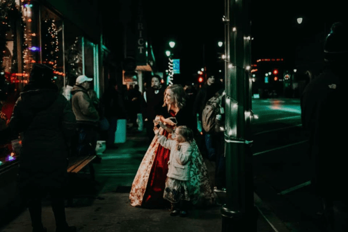 A woman in a red dress and a girl in a gray dress stand on a festive street, surrounded by holiday lights and people.