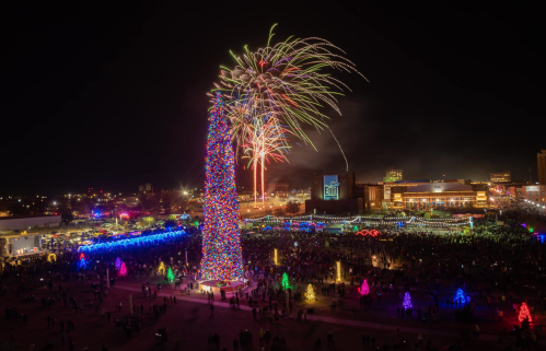A vibrant holiday celebration with a tall, lit Christmas tree and colorful fireworks over a crowd in a festive setting.
