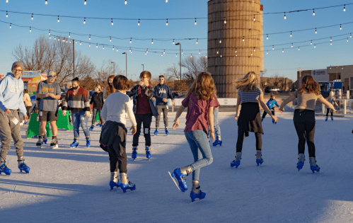 A lively outdoor ice skating rink with people skating and enjoying the evening under string lights.