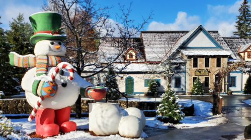 A large snowman statue with a candy cane stands in front of a snowy building, surrounded by evergreen trees.