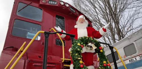 Santa Claus stands on a train engine, wearing a red suit and holding a festive wreath, surrounded by winter trees.