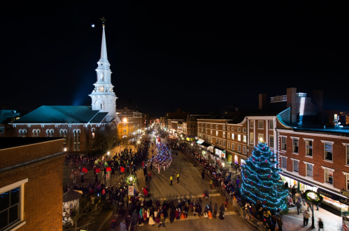 A festive night scene of a town square with a church steeple, decorated trees, and a crowd enjoying holiday lights.