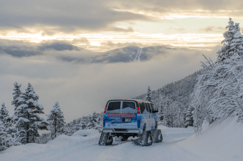 A blue vehicle with snow tracks drives through a snowy landscape, surrounded by mountains and clouds at sunset.