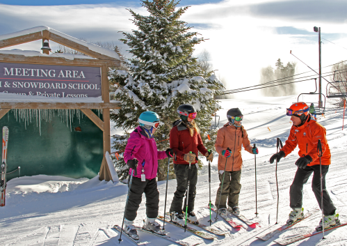 Four skiers in winter gear gather near a ski school sign, with snow-covered slopes and trees in the background.