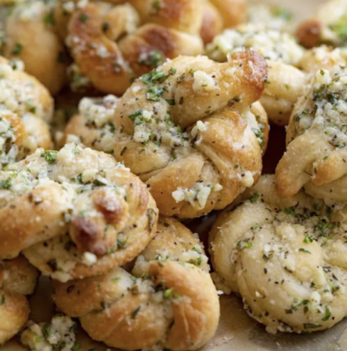 A close-up of golden, garlic herb knots topped with parsley and cheese, arranged on a baking sheet.