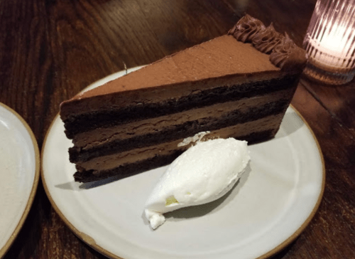 A slice of chocolate cake with layered frosting, served with a dollop of whipped cream on a plate.
