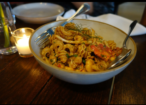 A bowl of pasta with shrimp, herbs, and a candle in the background on a wooden table.