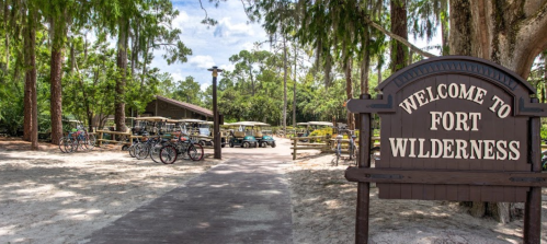 Sign reading "Welcome to Fort Wilderness" with a path leading to a wooded area and parked bicycles in the background.