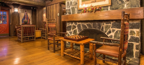 Cozy interior with wooden furniture, a stone fireplace, and a checkers table in a rustic setting. Warm lighting enhances ambiance.