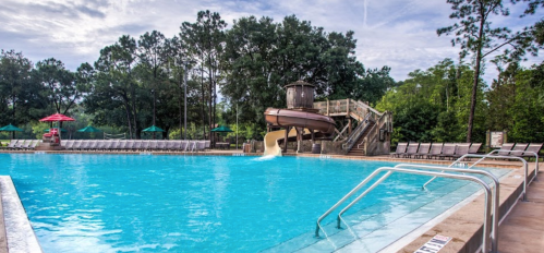 A clear blue pool with a water slide, surrounded by lounge chairs and green trees under a cloudy sky.