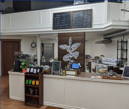 A cozy café interior featuring a counter with a menu board, coffee equipment, and a display of jars and plants.