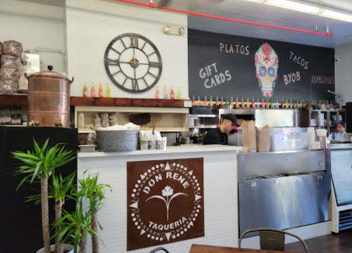 Interior of a taqueria with a wooden counter, clock, and colorful decor, featuring a menu and staff preparing food.