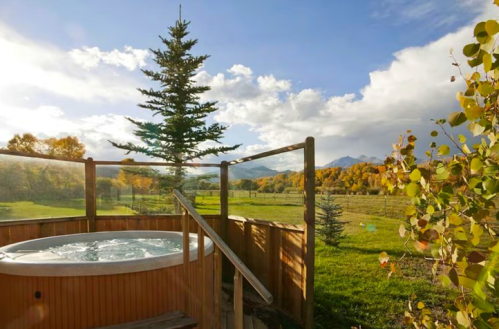 A wooden hot tub on a deck, surrounded by trees and a scenic view of mountains and autumn foliage.