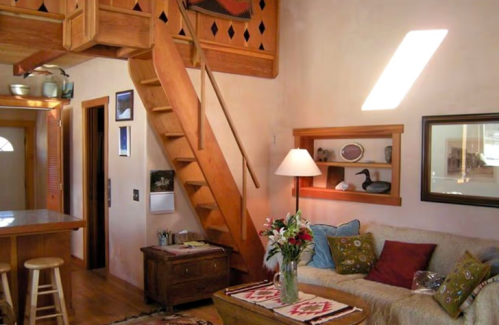 Cozy living room with wooden stairs, a sofa, a lamp, and decorative shelves, featuring warm tones and natural light.
