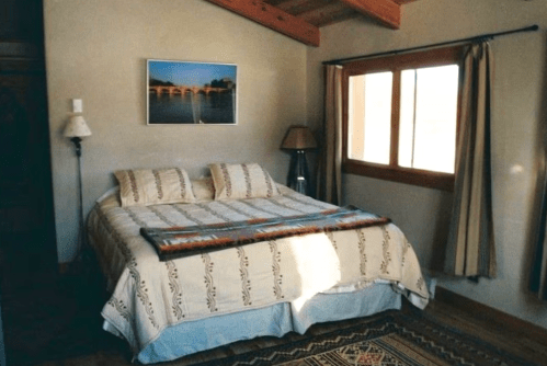 Cozy bedroom with a double bed, decorative blanket, lamp, and a window with curtains, featuring a scenic wall art.