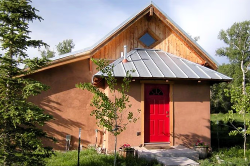 A small, rustic house with a red door, wooden accents, and a metal roof, surrounded by greenery.