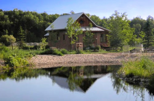 A serene landscape featuring a house by a pond, surrounded by trees and greenery under a clear blue sky.