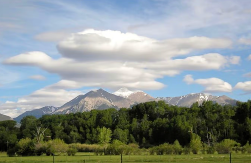 A scenic view of mountains with snow-capped peaks under a sky filled with fluffy clouds and green trees in the foreground.