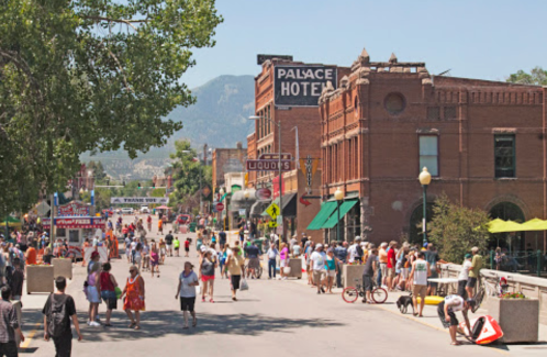 A busy street scene with people walking, shops, and mountains in the background on a sunny day.