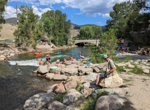 A scenic river scene with people kayaking, relaxing on rocks, and enjoying a sunny day by the water.