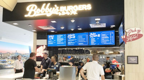 A busy burger restaurant counter with staff, a menu board, and customers in a modern setting.