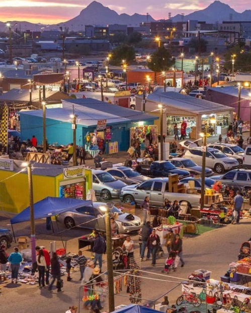 A bustling outdoor market at sunset, with colorful stalls, shoppers, and parked cars in a lively atmosphere.