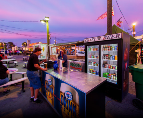 A vendor at a beer stand during sunset, with colorful skies and people in the background enjoying the fair atmosphere.