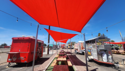 Colorful food trucks lined up under red shade sails, with wooden picnic tables in a sunny outdoor setting.