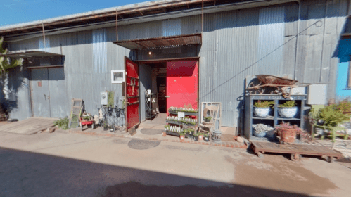 A rustic storefront with red doors, displaying plants and decor outside, set against a corrugated metal building.