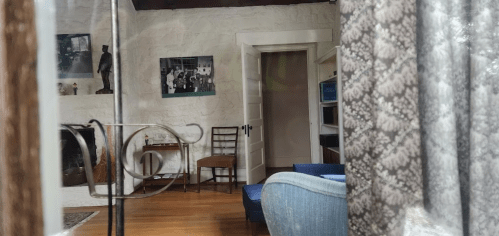 A cozy room with a fireplace, wooden floor, and a blue chair, featuring framed photos on the wall and a door leading outside.