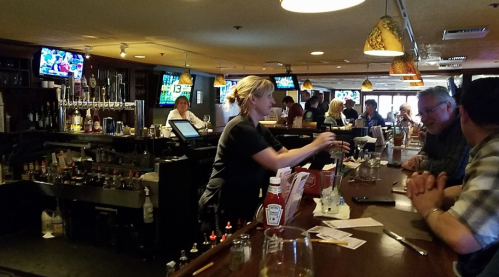 A busy bar scene with a bartender serving drinks, patrons at the counter, and TVs showing sports in the background.