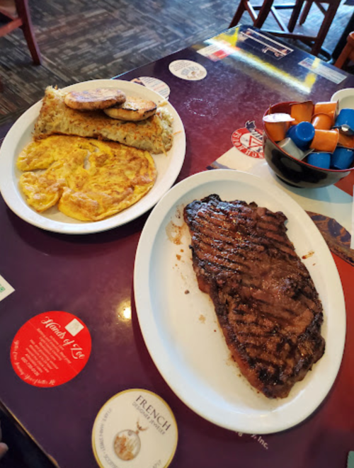 A plate with a grilled steak, an omelet, and hash browns, alongside a bowl of colorful condiments on a restaurant table.