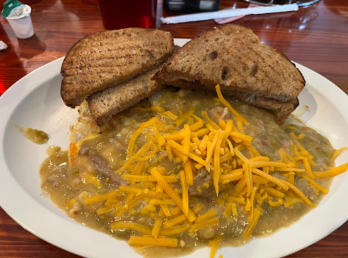 A plate of green chili topped with shredded cheese, served with two slices of toasted bread on the side.