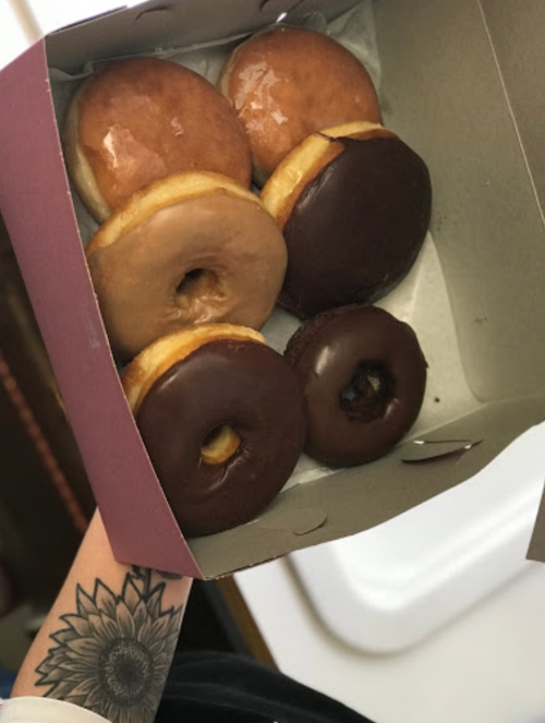 A hand holding a box of assorted donuts, including glazed and chocolate-covered varieties.