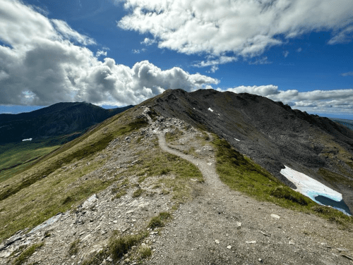 A winding trail leads up a rocky mountain ridge under a partly cloudy sky, with green hills in the background.