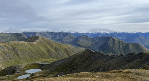 A panoramic view of rugged mountains with patches of grass and small lakes under a cloudy sky.