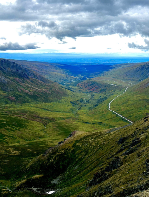 A panoramic view of a lush green valley surrounded by mountains under a cloudy sky. A winding road cuts through the landscape.