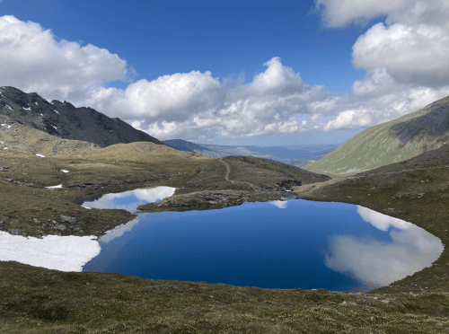 A serene mountain landscape featuring a clear blue lake reflecting clouds and surrounding hills under a bright sky.