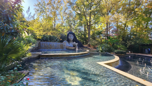 A serene garden scene featuring a large statue of a woman beside a tranquil water feature surrounded by lush greenery.