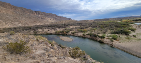 A winding river flows through a desert landscape, surrounded by mountains and sparse vegetation under a cloudy sky.